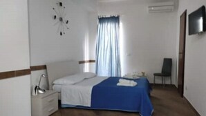 Un dormitori amb un llit, una tauleta de nit, una cadira, una televisió i una finestra amb cortines.