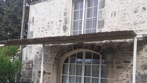 Un edificio rústico de piedra con tejado de tejas, un gran ventanal arqueado y un patio cubierto con mesa y sillas.