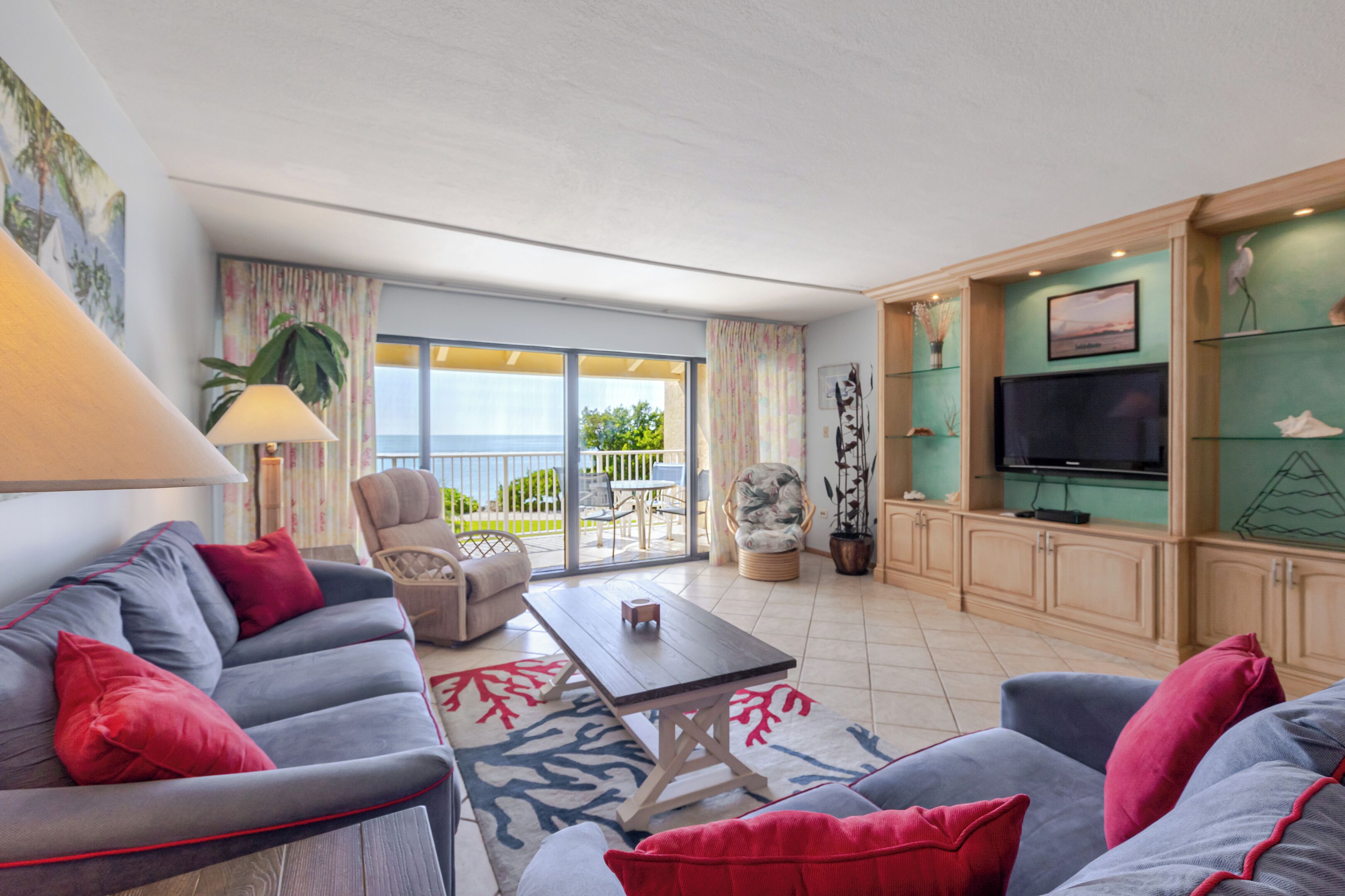 A modern living room with a sofa, armchair, and a dining table. There is a TV on a cabinet, a plant, and a view of the outdoors through a sliding door.