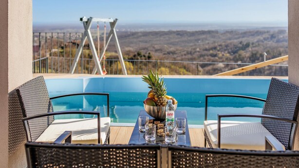 Una mesa junto a la piscina con dos sillas de mimbre, un cuenco de fruta y una botella de Jana.