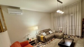 Ein Wohnzimmer mit Ecksofa, Couchtisch, einer Lampe und einer Stehlampe. Im Hintergrund ist ein großes Fenster mit Vorhängen und ein Bücherregal zu sehen.