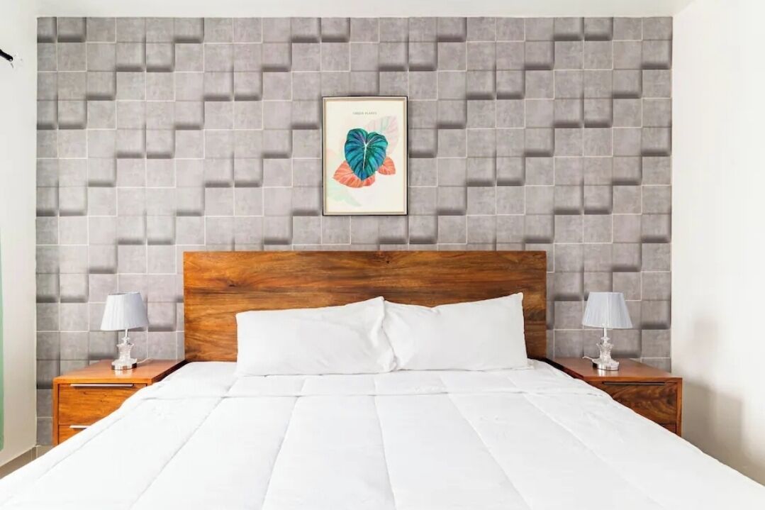 Ein Hotelzimmer mit einem hölzernen Kopfteil, einem großen Bett mit weißer Bettwäsche, zwei Nachttischen mit Lampen und einer grauen Wand mit Muster, über dem Bett befindet sich ein gerahmtes Kunstwerk.