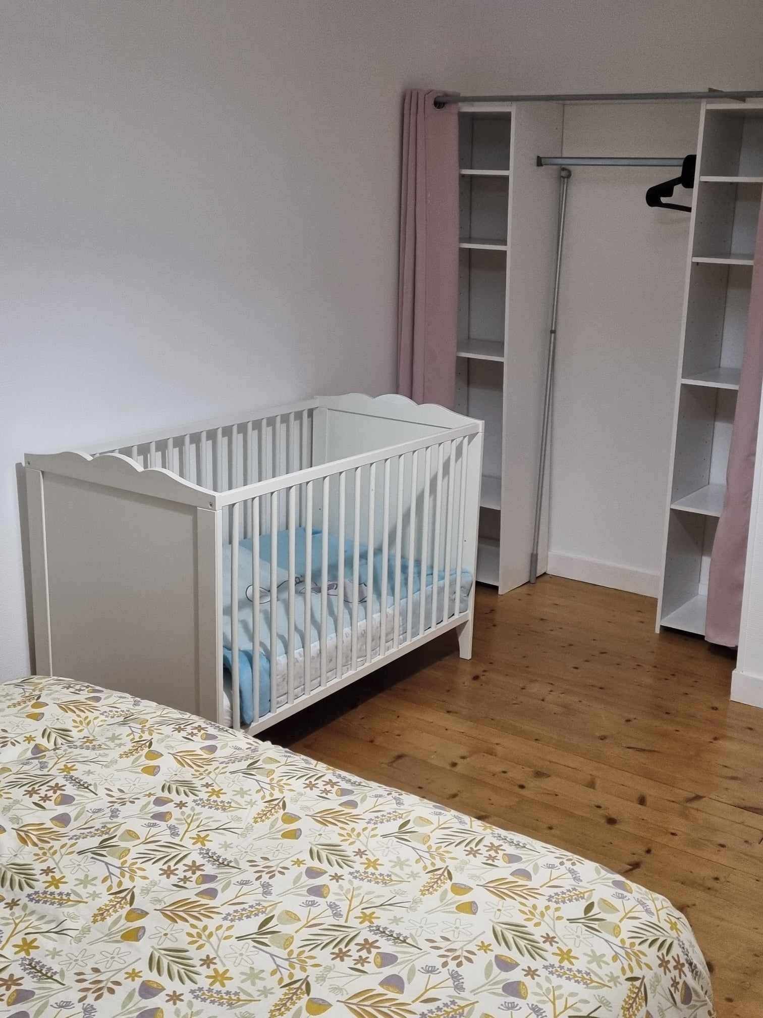 Una habitación con una cuna blanca, una cama con edredón floral, suelo de madera y un armario blanco con cortinas rosadas.