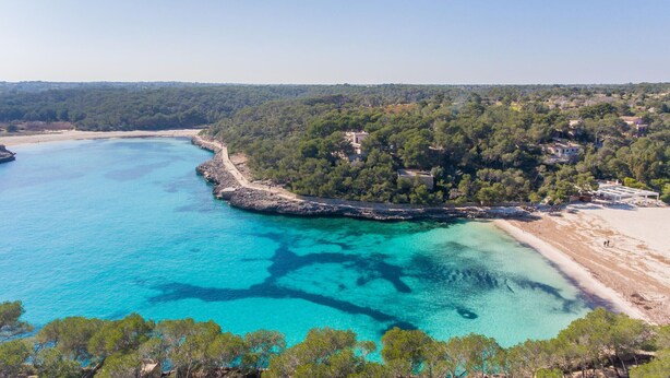 Luftaufnahme einer Küstenlandschaft mit klarem türkisblauem Wasser, einer felsigen Küstenlinie und einer dicht bewaldeten Halbinsel mit Gebäuden.