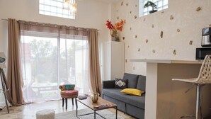 A modern living room with a sofa, coffee table, and a small stool. There is a kitchen area with a microwave and a chair. - Appartement Proche de Disney (Chalifert)