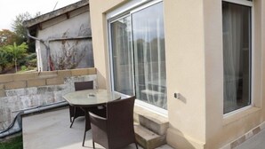 A patio with a glass table and wicker chairs, a sliding glass door, and a concrete wall with a drainage pipe. - Appartement Proche de Disney (Chalifert)