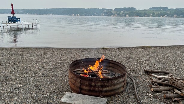 Een kiezelstrand met een metalen vuurkuil en houtblokken, een steiger met stoelen en een rustig meer met gebouwen in de verte.