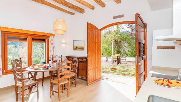 A dining area with wooden chairs and a table, a kitchen with a sink, and an open door leading to an outdoor area with a bench and a tree. - Masia Can Casas (Sant Iscle de Vallalta)