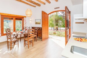 A dining area with wooden chairs and a table, a kitchen with a sink, and an open door leading to an outdoor area with a bench and a tree. - Masia Can Casas (Sant Iscle de Vallalta)
