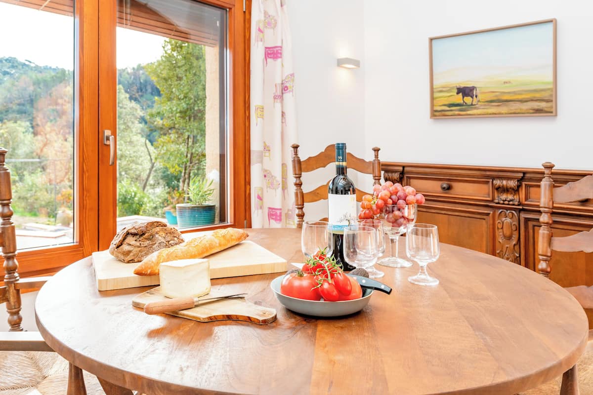 A round wooden dining table with a rustic bread, cheese, tomatoes, and wine glasses.