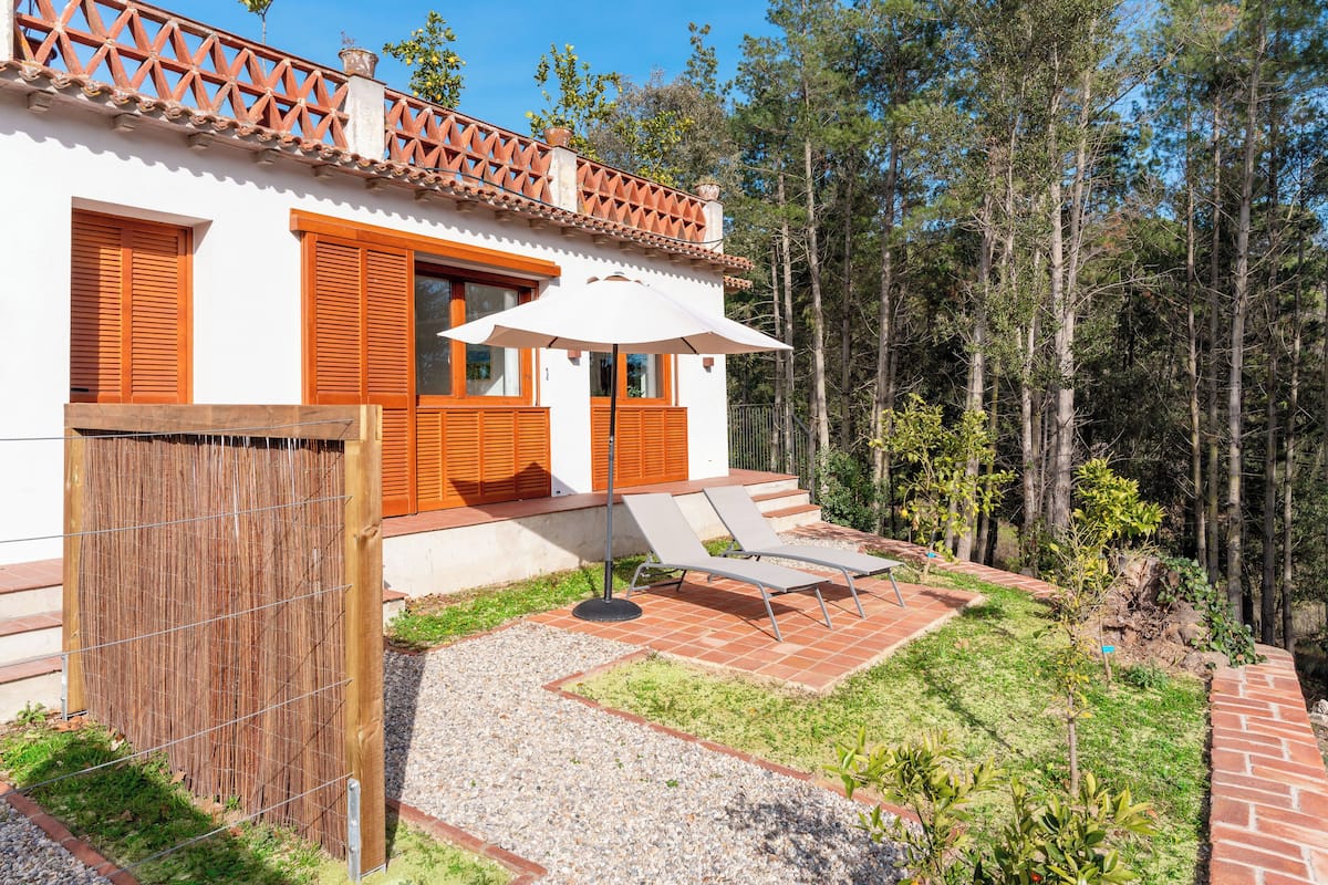 A white building with orange shutters, a wooden fence, and a patio with two sun loungers and a table.