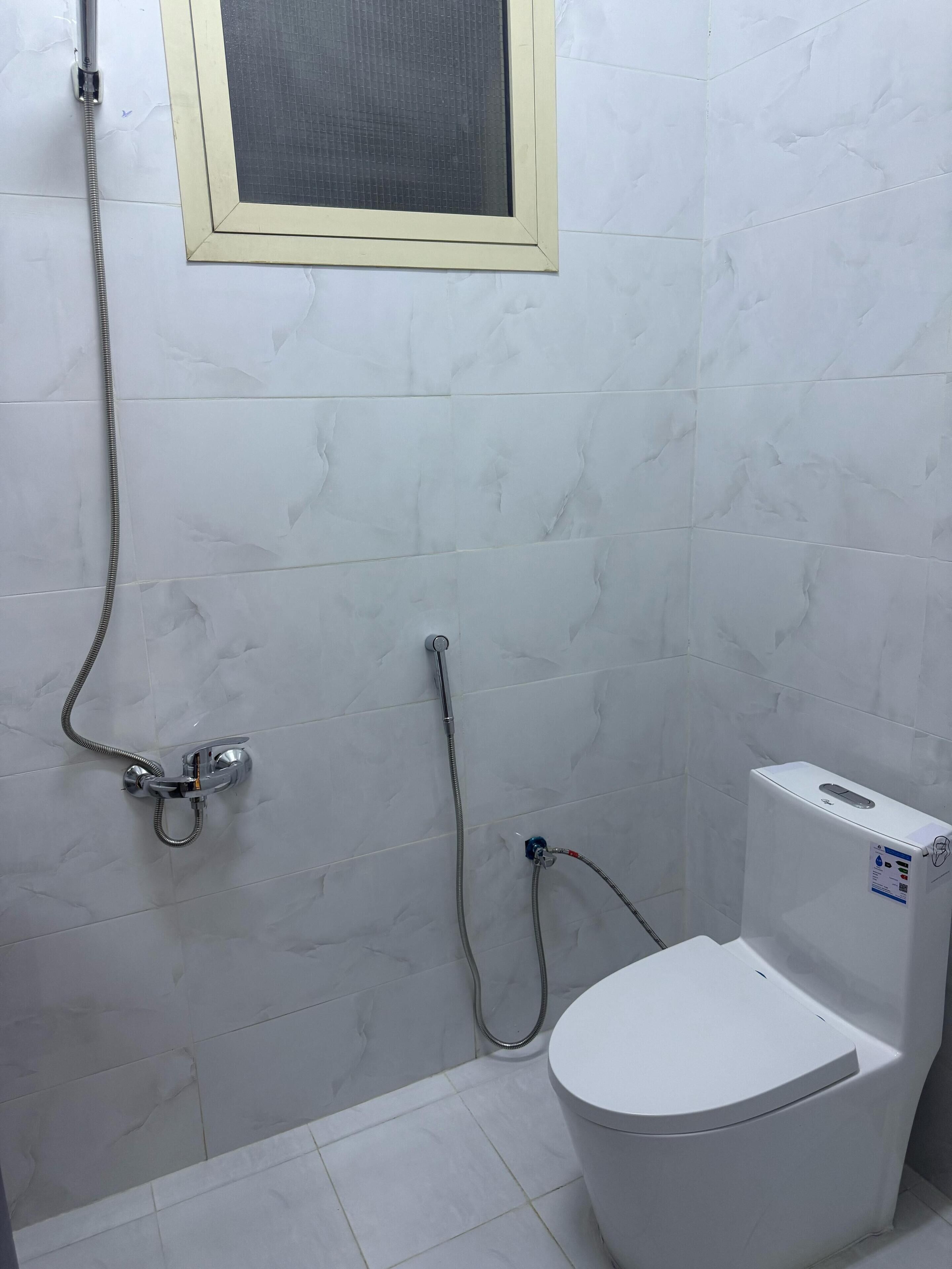Ein Badezimmer mit weißen Marmorfliesen, einer Dusche inklusive Handbrause und einer Toilette.