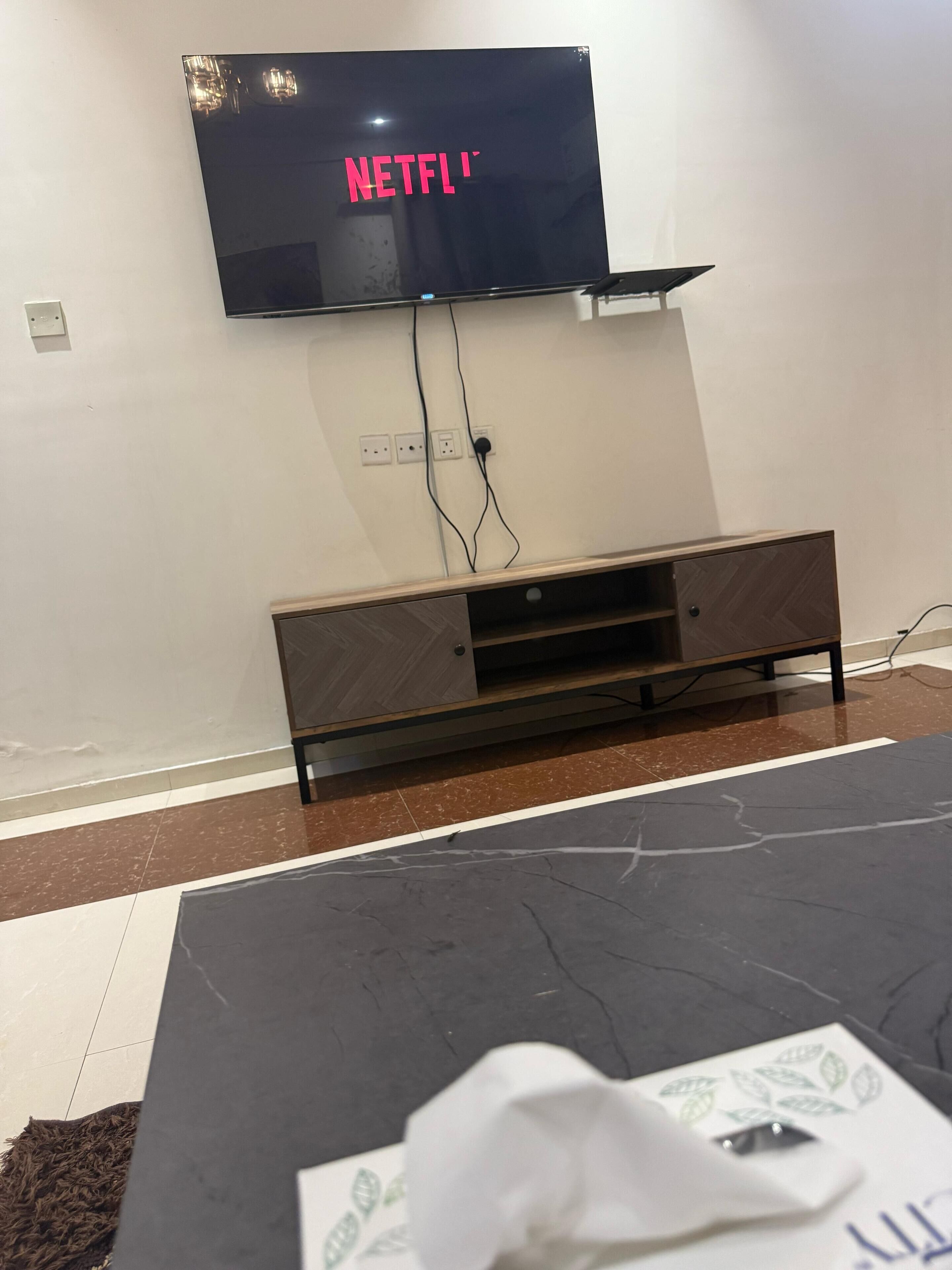 Une pièce dotée d’un téléviseur à écran plat fixé au mur, sur lequel est affiché le logo de Netflix, d’un meuble de rangement en bois en dessous et d’un tapis à motifs sur le sol.