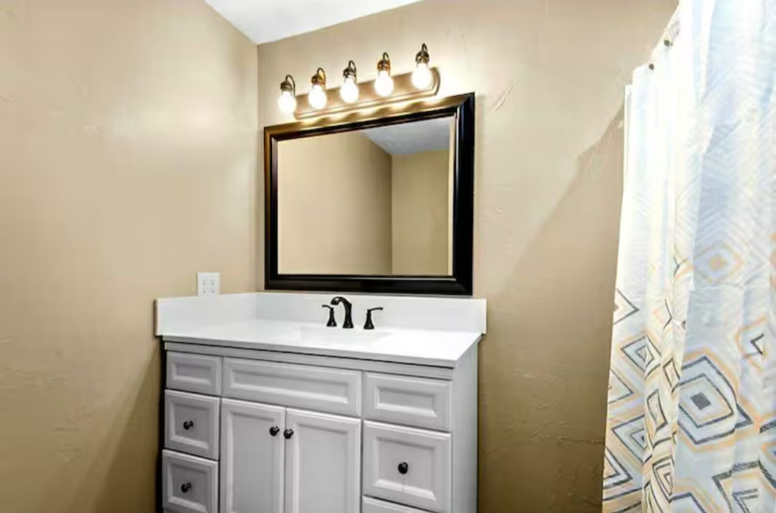 A bathroom with a white vanity, a large mirror with lights, and a patterned shower curtain.