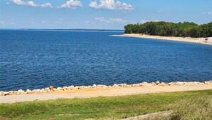 A coastal landscape with a sandy beach, a grassy foreground, and a forested area. - Lake Front Cabin! 
Pool, Basketball, Boat parking & Firepit. Lots of parking. (Brookeland)