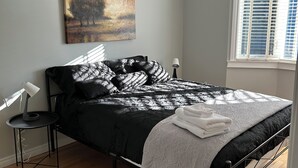 Ein Schlafzimmer mit Bett, Nachttisch, Lampe, einem Gemälde und einem Fenster mit Jalousien.