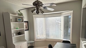 Ein Essbereich mit Tisch und Stühlen, einem Deckenventilator, einem Fenster mit Jalousien und einem weißen Schrank mit Regalen.