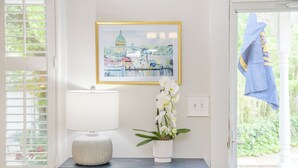 A dark wooden console table with a white lamp and a white vase with orchids. A framed painting of a cityscape hangs above the table. - Historic Annapolis Home! (Annapolis)