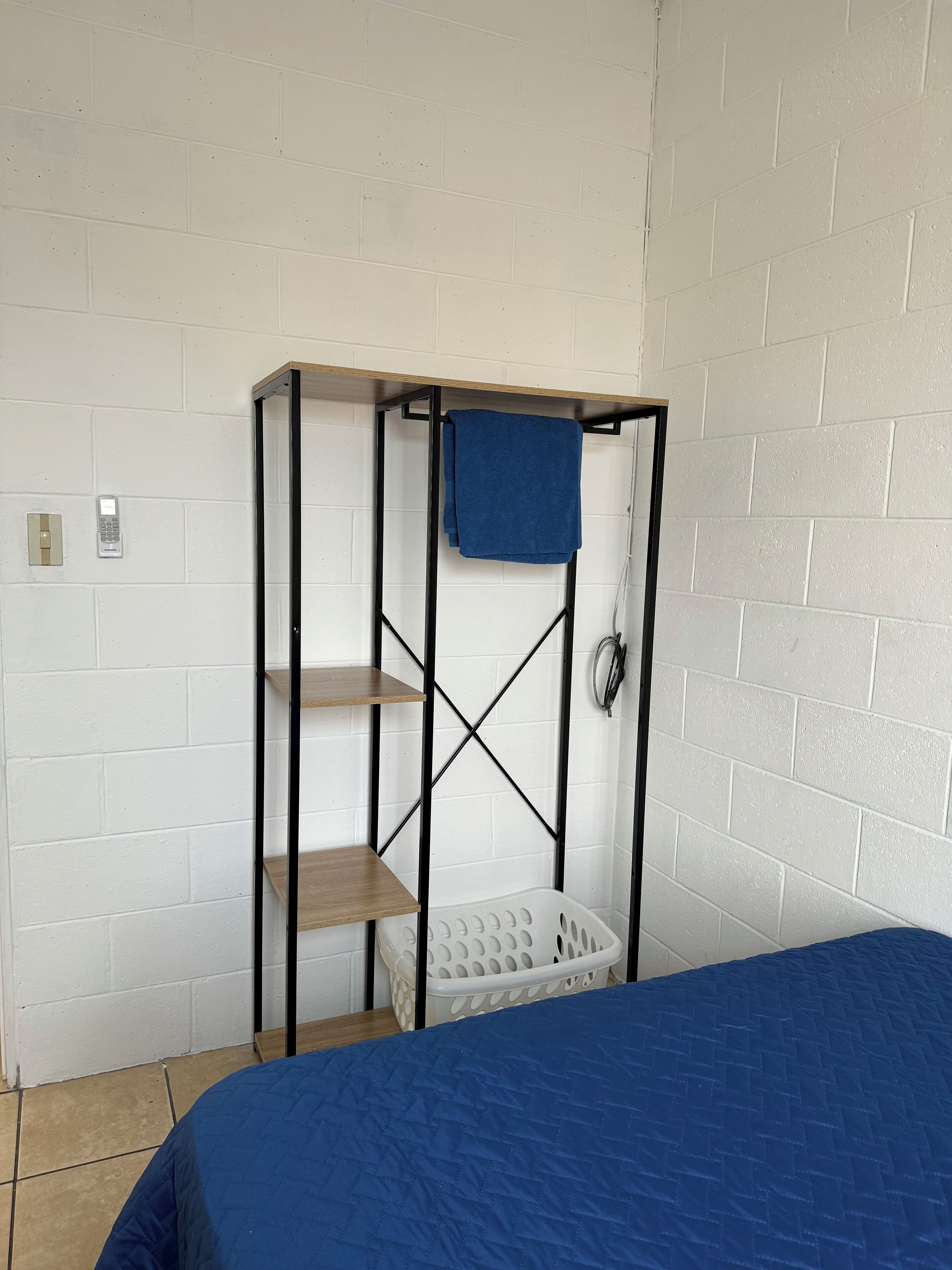 Una habitación con cama, un armario metálico con estantes, una cesta para ropa colgada y un teléfono fijo en la pared.