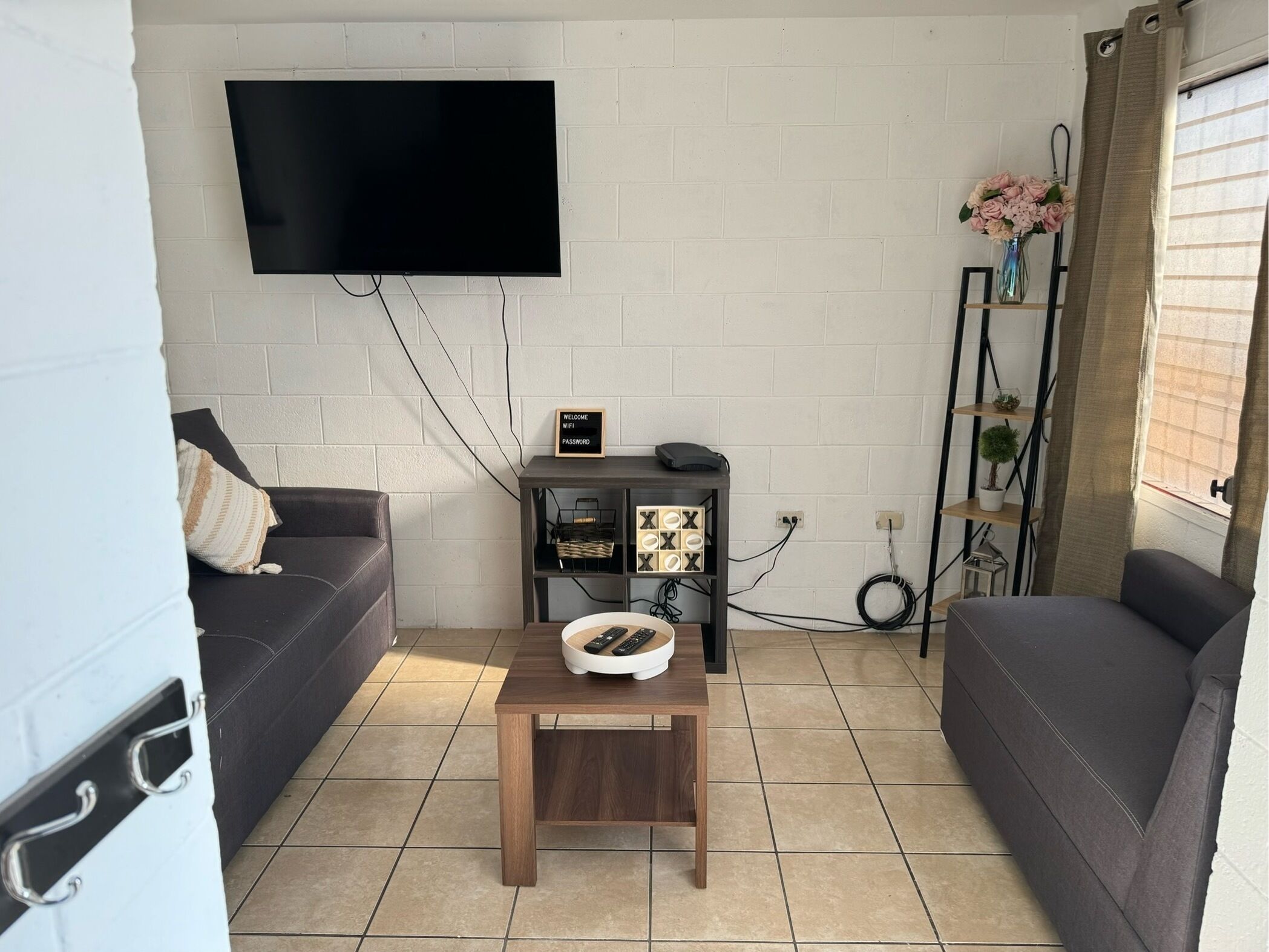 Ett vardagsrum med en platt-TV som är monterad på en vit tegelvägg, ett litet träbord med en skål och fjärrkontroller, en mörkfärgad soffa och en hylla med växter och en välkomstskylt.