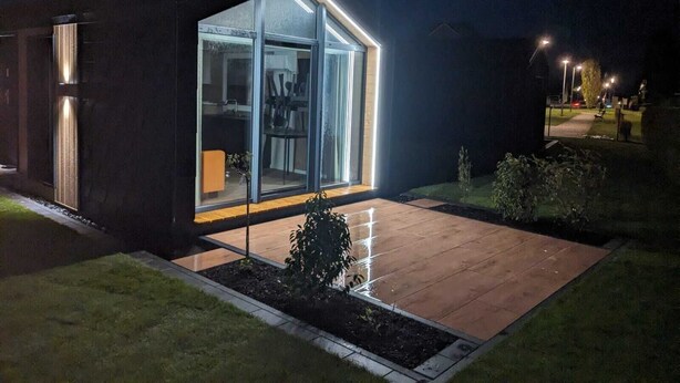 Una casa moderna amb façana de vidre, una terrassa de fusta i un jardí ben cuidat.