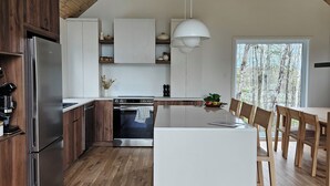 A modern kitchen with wooden flooring, a central island, and a large window overlooking a forest.