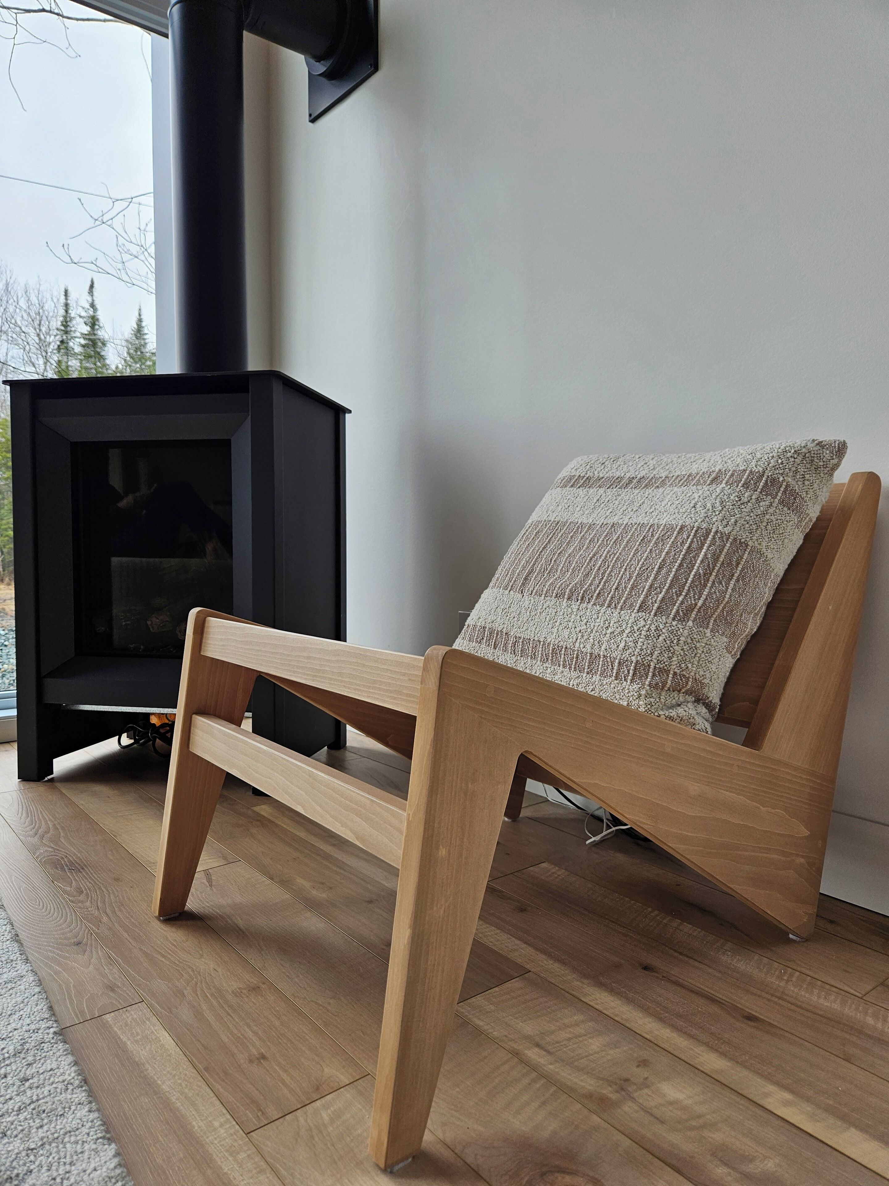 En modern trästol med en vävd prydnadskudde, placerad nära en svart vedeldad kamin i ett rum med trägolv och en vit vägg.