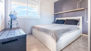 Ein Schlafzimmer mit Bett, Standventilator, Fenster mit Jalousien und Nachttisch.