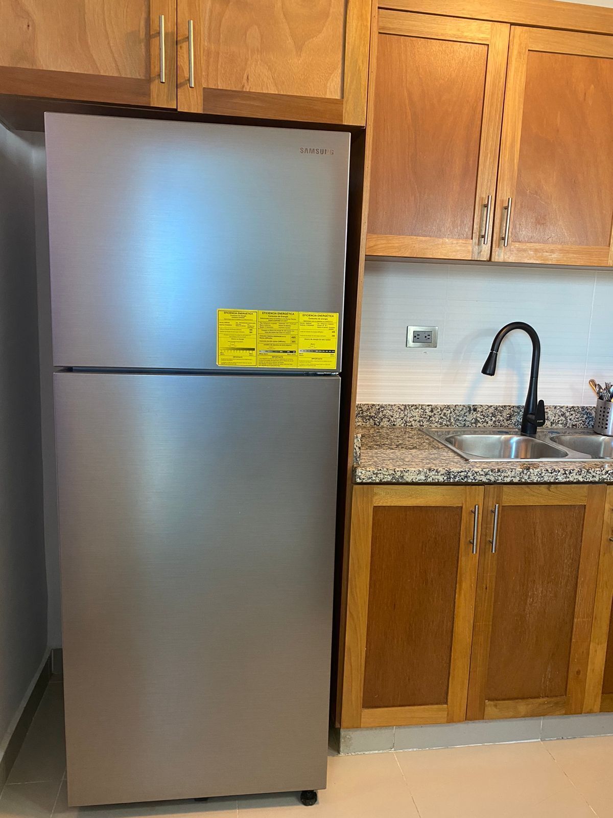 Ein Edelstahl-Kühlschrank mit einem gelben Aufkleber, Holzschränke und eine Arbeitsplatte aus Granit mit einem schwarzen Wasserhahn.