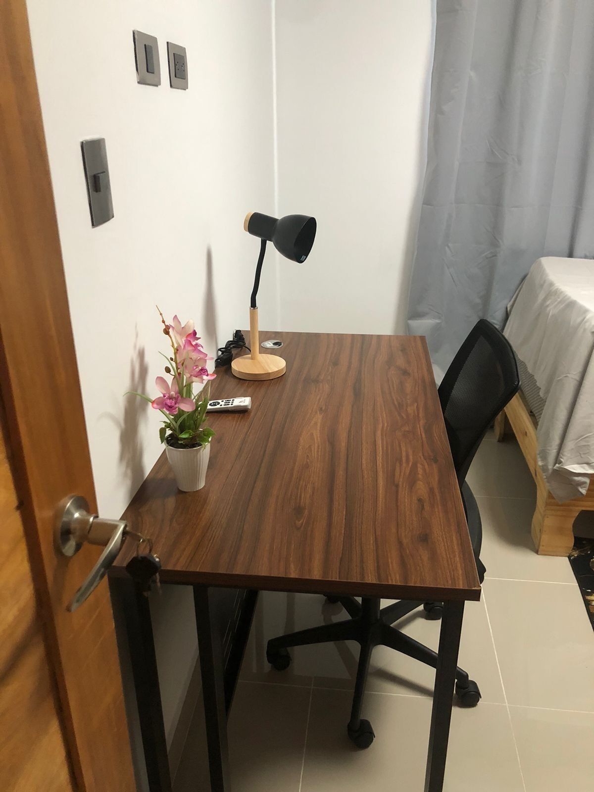 Una stanza piccola e ordinata con una scrivania in legno, una sedia da ufficio nera, una pianta in vaso e una lampada da tavolo.