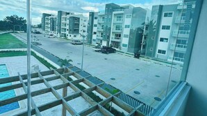 Un immeuble d’appartements moderne, doté de plusieurs étages et de balcons, un espace de stationnement avec des voitures, et un ciel bleu dégagé.