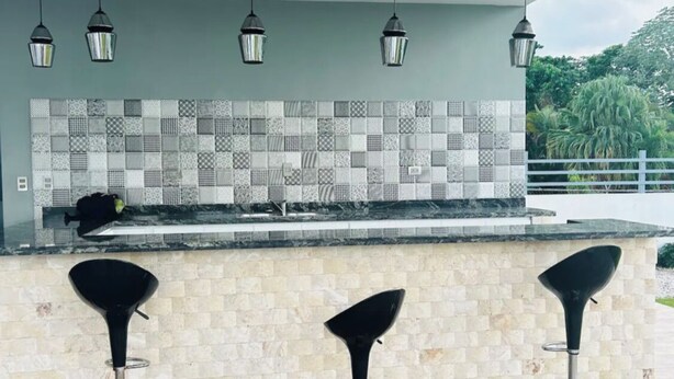 Une cuisine moderne avec un mur recouvert de carreaux, trois tabourets de bar noirs et des luminaires suspendus.