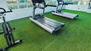 Une salle de sport dotée de tapis de course et d’appareils d’exercice, avec un sol vert, de grandes fenêtres et un ventilateur au plafond.