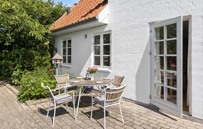 A white brick house with a tiled roof, a patio with a round wooden table and four wicker chairs, and a window with a view of the garden. - Nice home in Sønderborg with kitchen (Sønderborg)