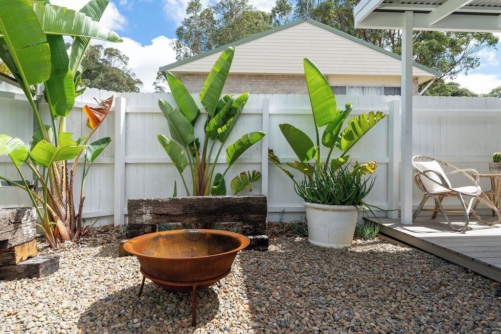 A backyard with a white fence, a wooden bench, a metal planter, and a wicker chair.