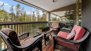 A covered outdoor balcony with wicker furniture, a small table with a plant, and a view of a forest. - Sj2503-3 (Keystone)