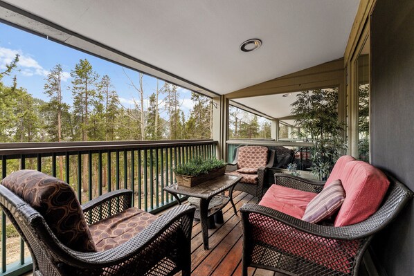 A covered outdoor balcony with wicker furniture, a small table with a plant, and a view of a forest. - Sj2503-3 (Keystone)