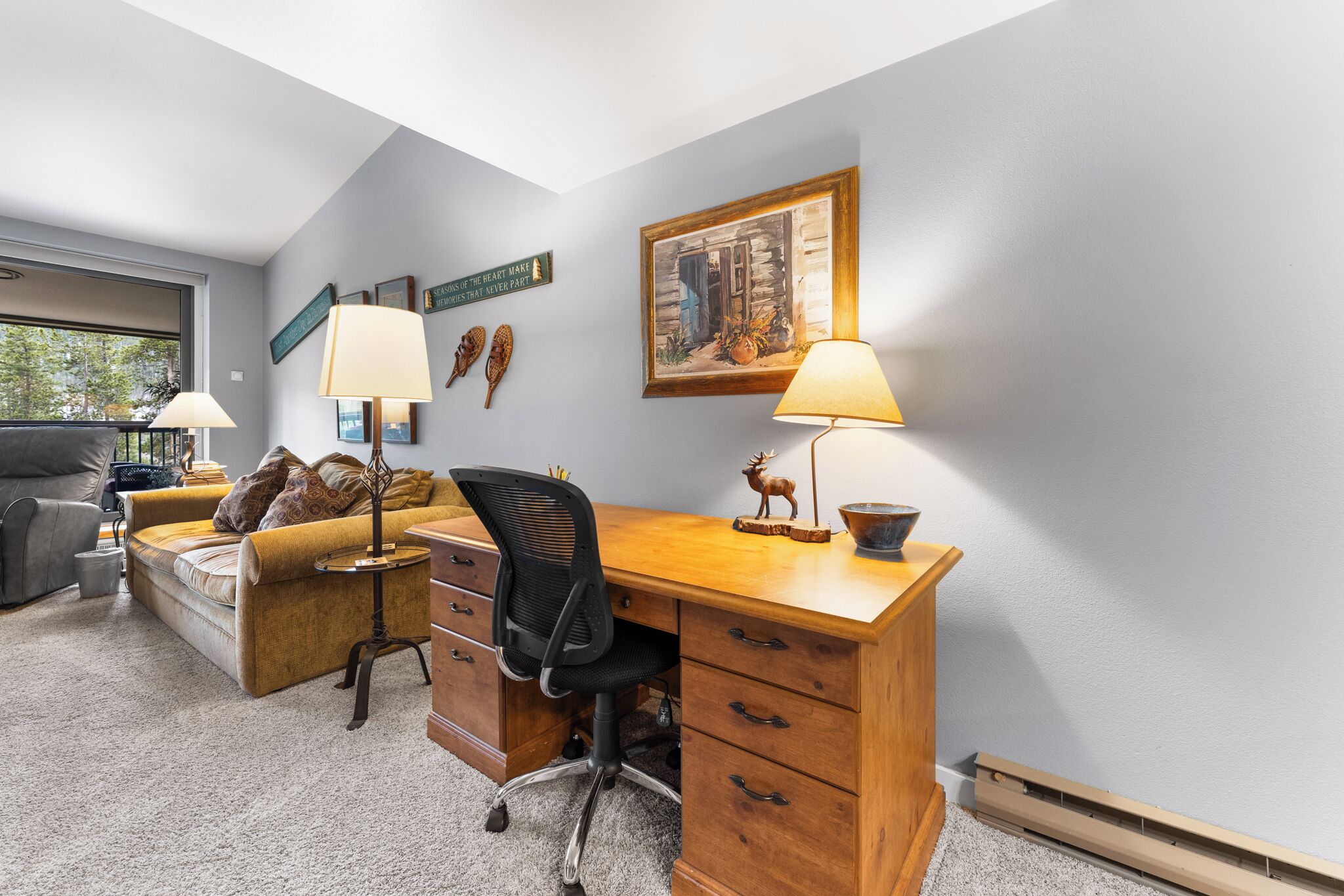 A home office with a wooden desk, two lamps, a chair, a couch, and a window.