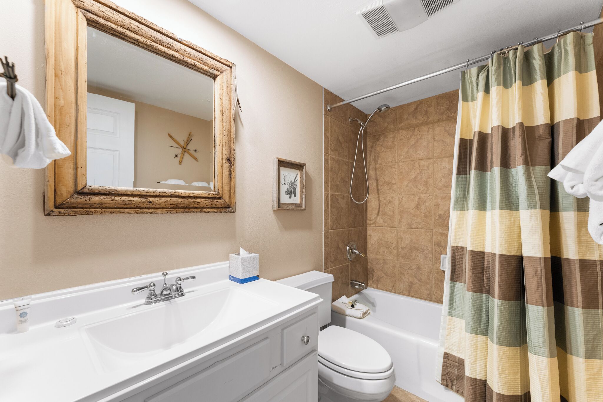 A bathroom with a white sink, toilet, and shower. There is a wooden-framed mirror above the sink, a towel hanging on the wall, and a shower curtain with a plaid pattern.
