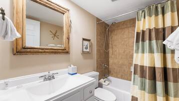 A bathroom with a white sink, toilet, and shower. There is a wooden-framed mirror above the sink, a towel hanging on the wall, and a shower curtain with a plaid pattern.