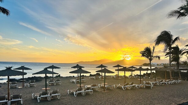 Ein Strand mit Liegestühlen, strohgedeckten Sonnenschirmen, felsiger Küste und einem Sonnenuntergang im Hintergrund.