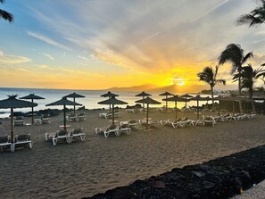 Una playa con filas de tumbonas y sombrillas de paja, una costa rocosa y un atardecer de fondo.