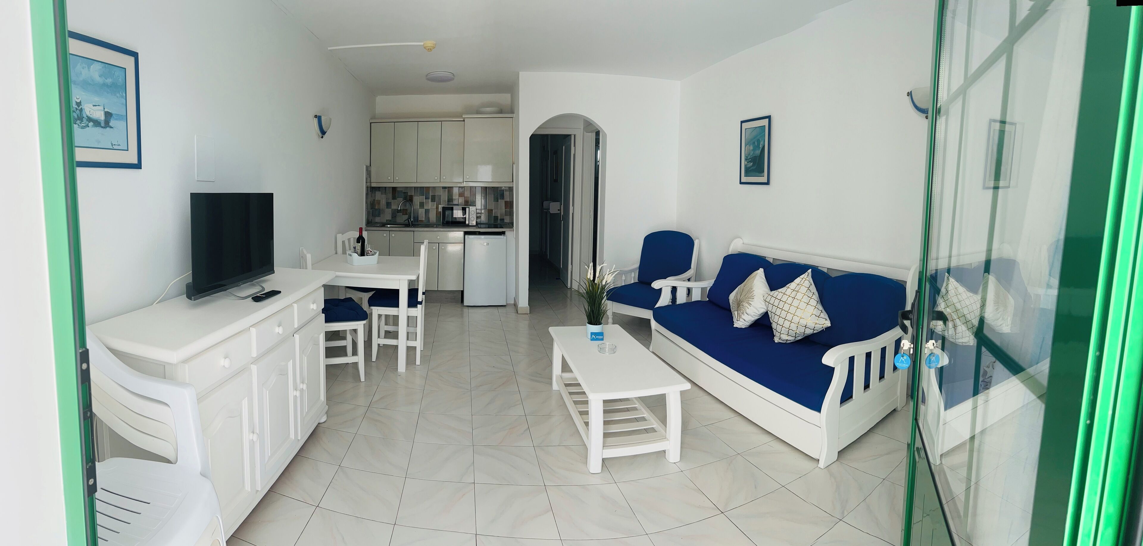 Una sala de estar moderna con televisor, zona de estar y una cocina al fondo.