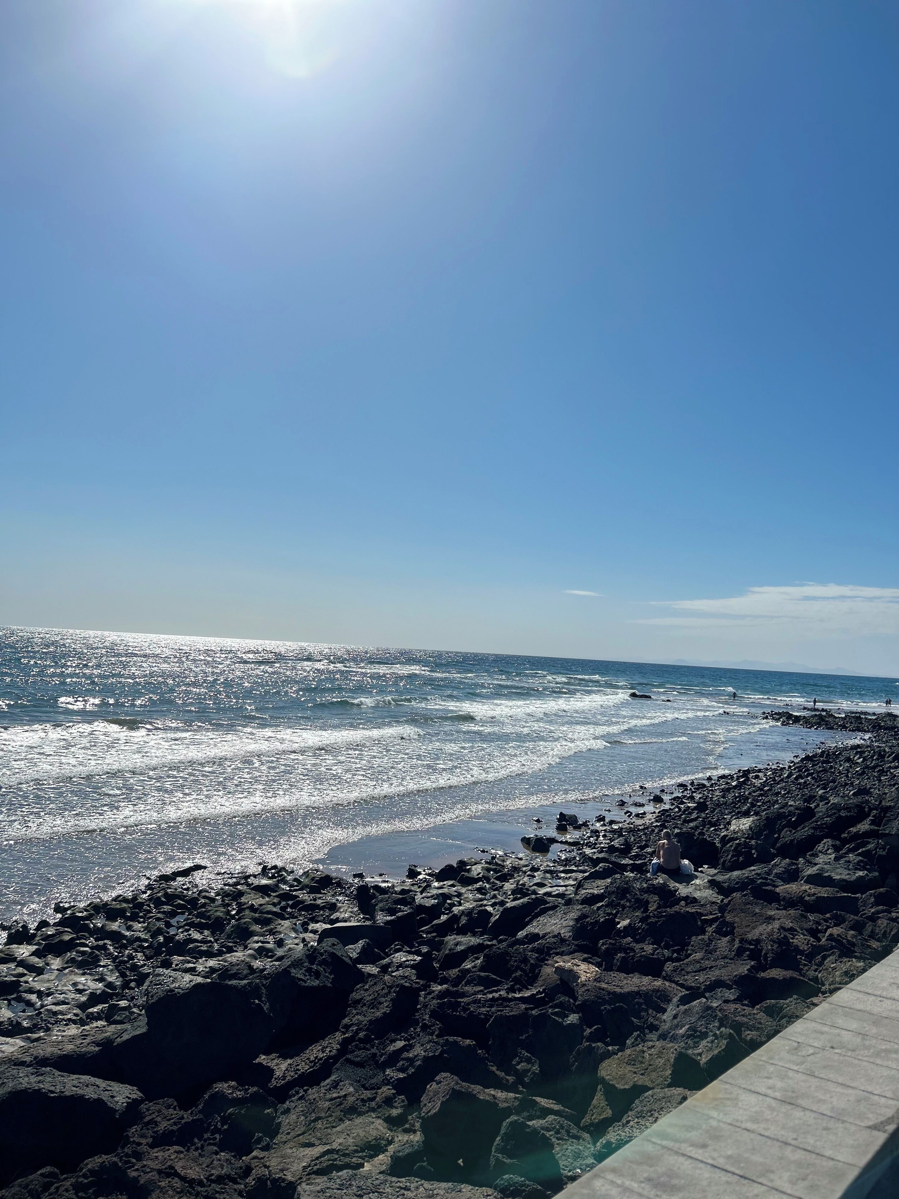 Une côte rocheuse, avec des vagues, un ciel dégagé et des montagnes à l’horizon.