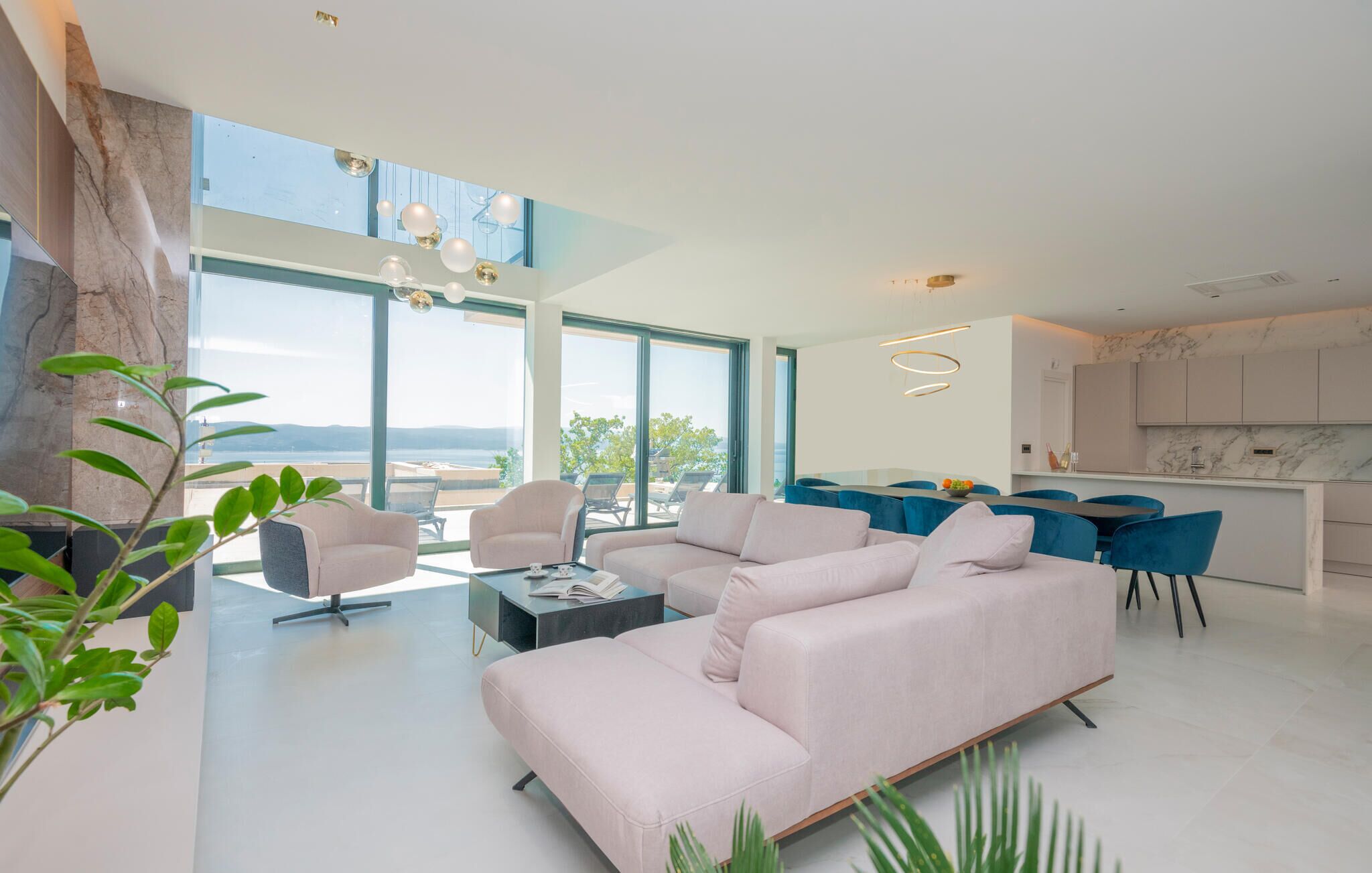 En moderne stue med en stor sofa, to lænestole, et sofabord og et spiseområde med blå stole. Der er et stort vindue med udsigt til naturen.