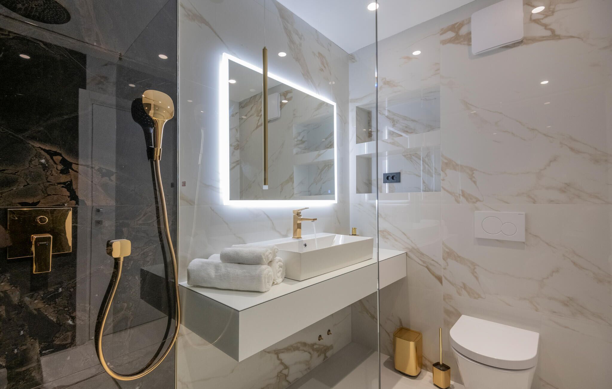 Un baño moderno con ducha de vidrio, lavamanos blanco y detalles dorados, y pared de mármol.