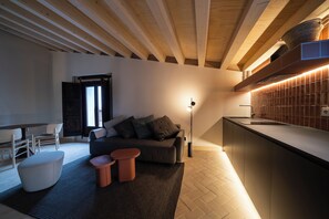 Ein modernes Interieur mit einer Couch, Hocker und einem kleinen Tisch in einem Wohnbereich. Eine Küche mit Arbeitsplatte und Schränken ist ebenfalls zu sehen.