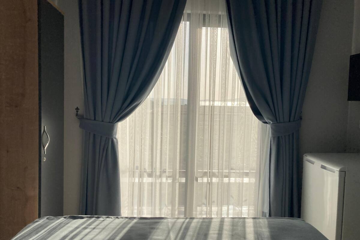Un dormitorio con cama, ventana con cortinas traslúcidas y radiador.