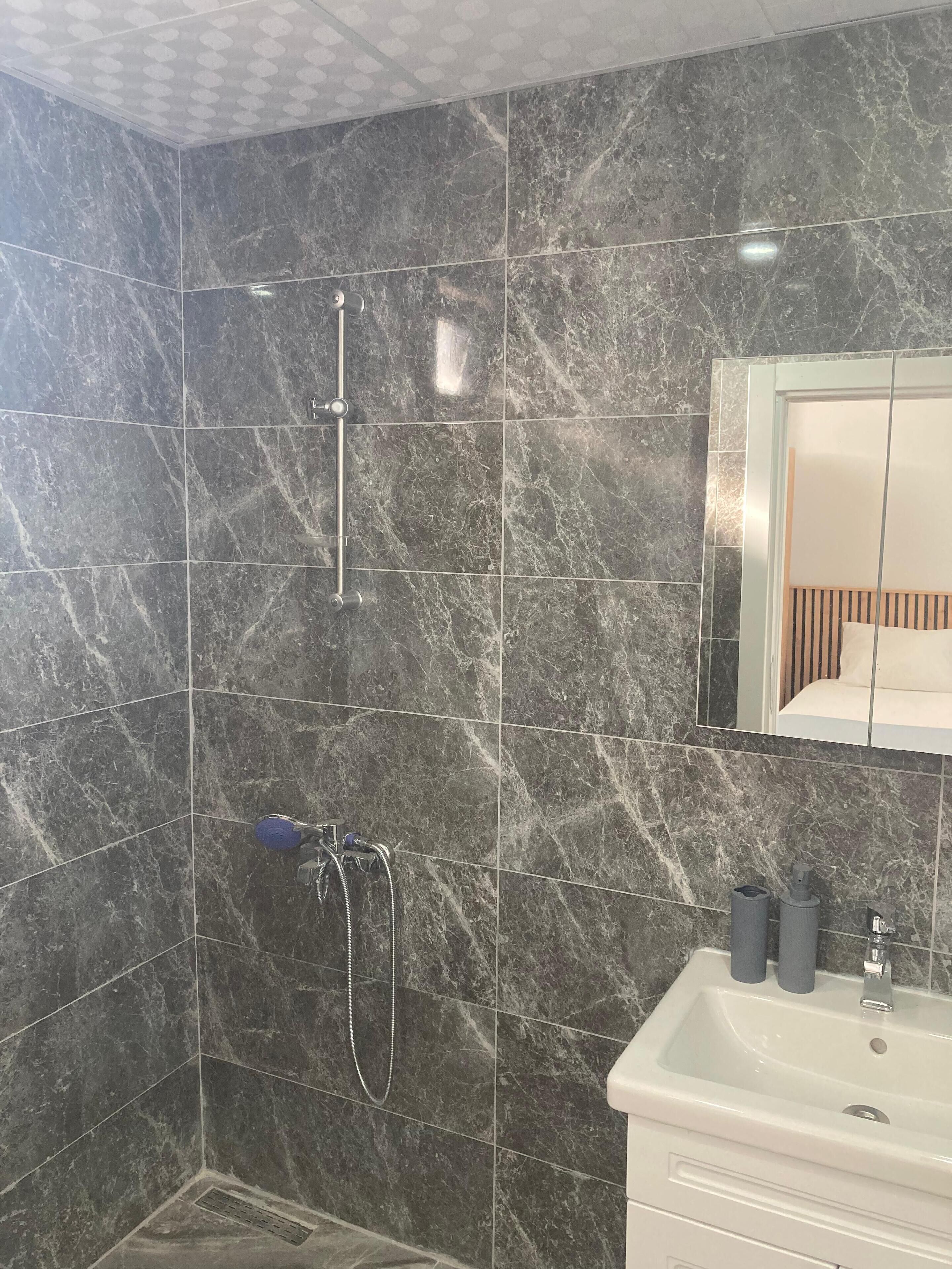 Ein Badezimmer mit dunklen Marmorfliesen, einer Dusche, einem Waschbecken mit Spiegel und Toilettenartikeln.