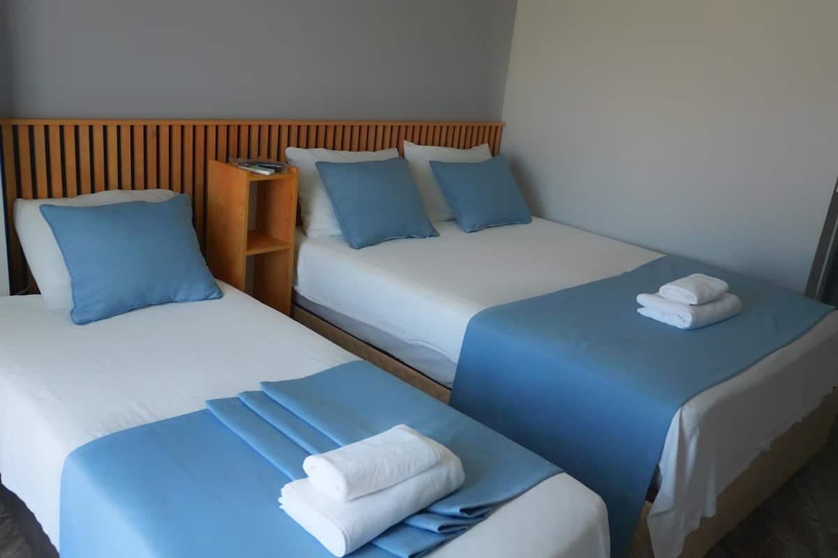 Habitación de hotel con dos camas, cada una con cubrecama azul y almohadas blancas. Se observa un cabecero de madera, una mesita de noche con lámpara y una toalla doblada sobre la cama de la derecha.